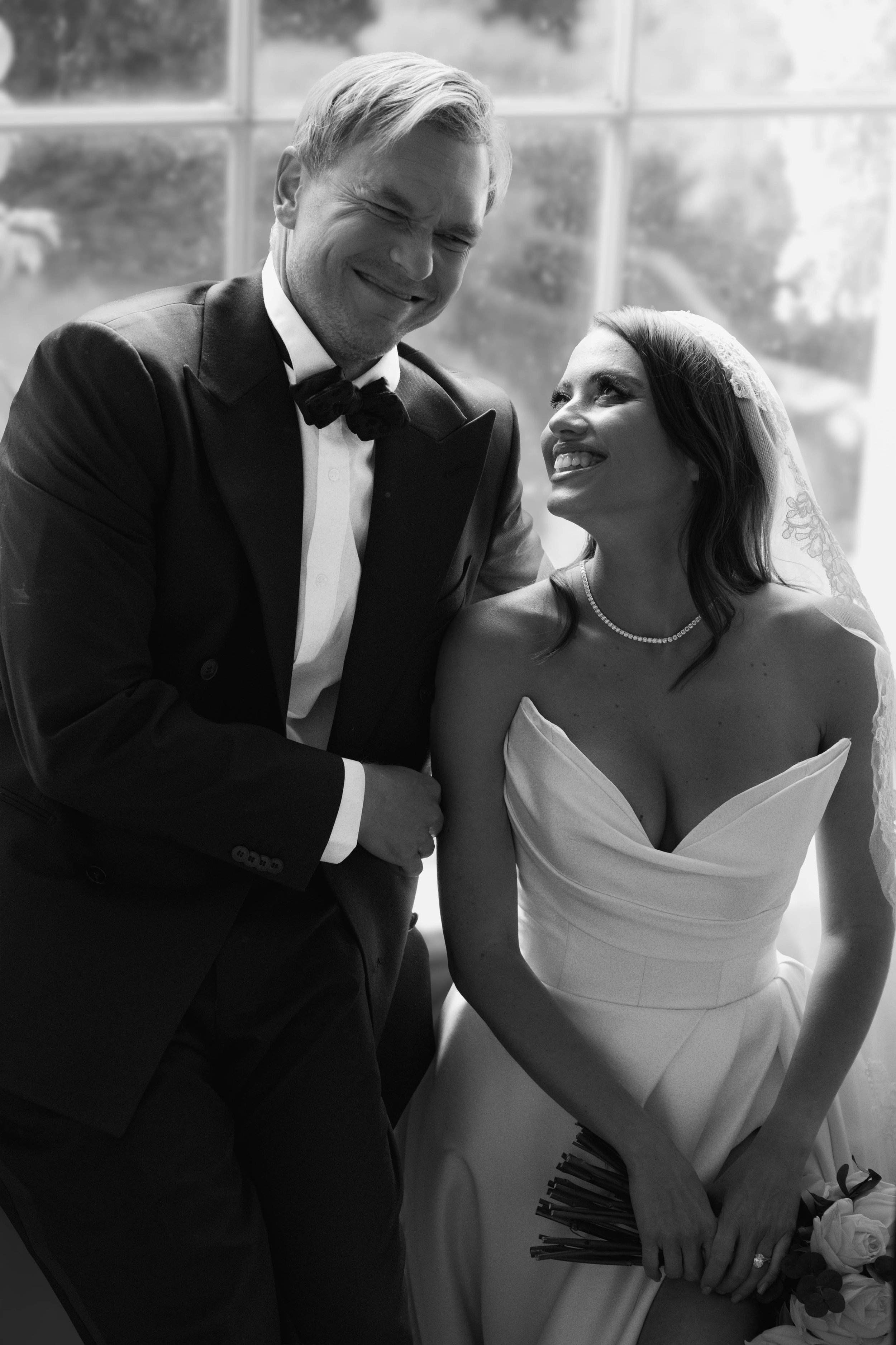 A black and white photo of a smiling groom in a tuxedo leaning towards a smiling bride in a wedding dress, holding a bouquet of roses.