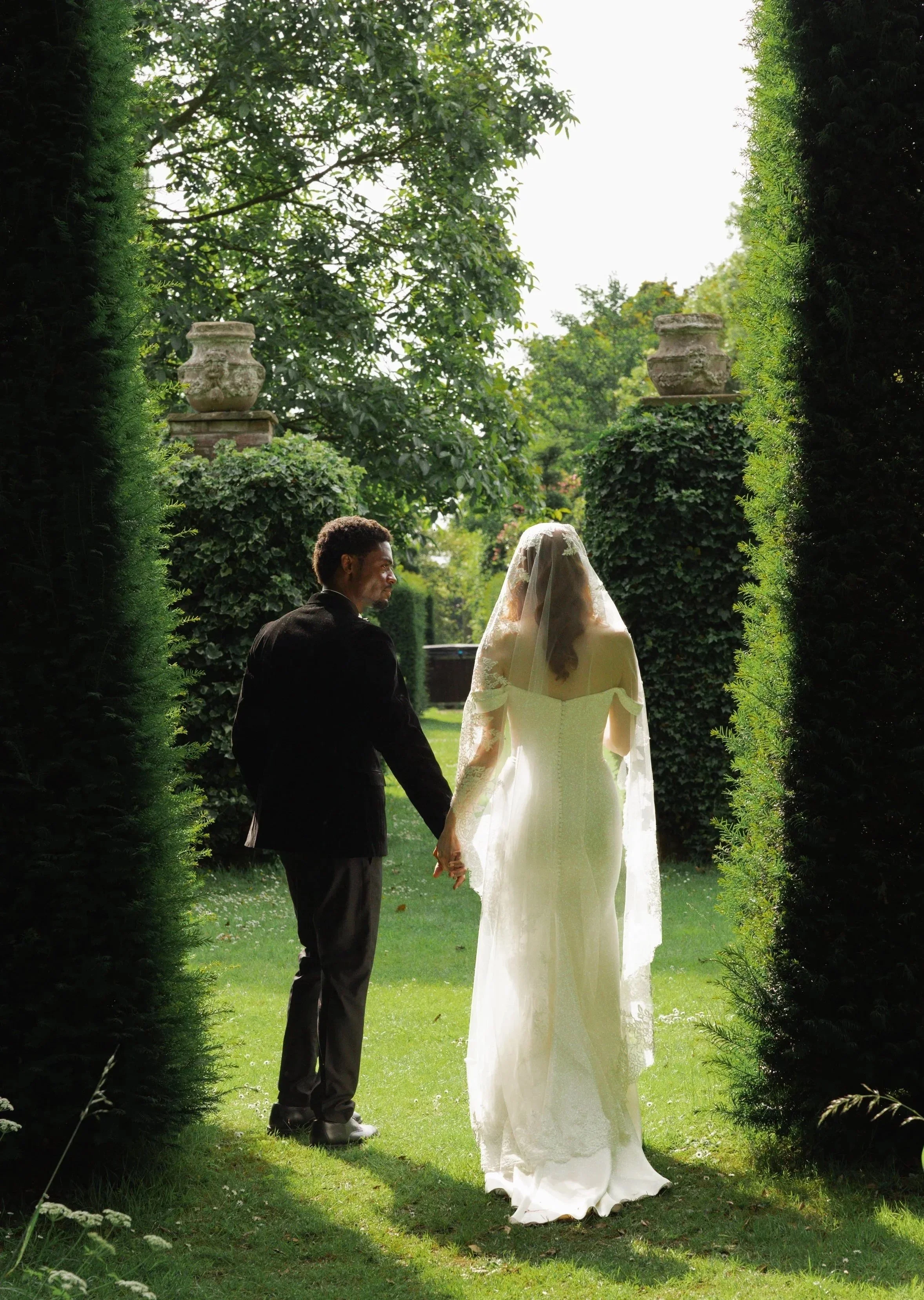 A couple holding hands and facing each other outdoors, in a garden with trees and greenery, with statues on pillars in the background.