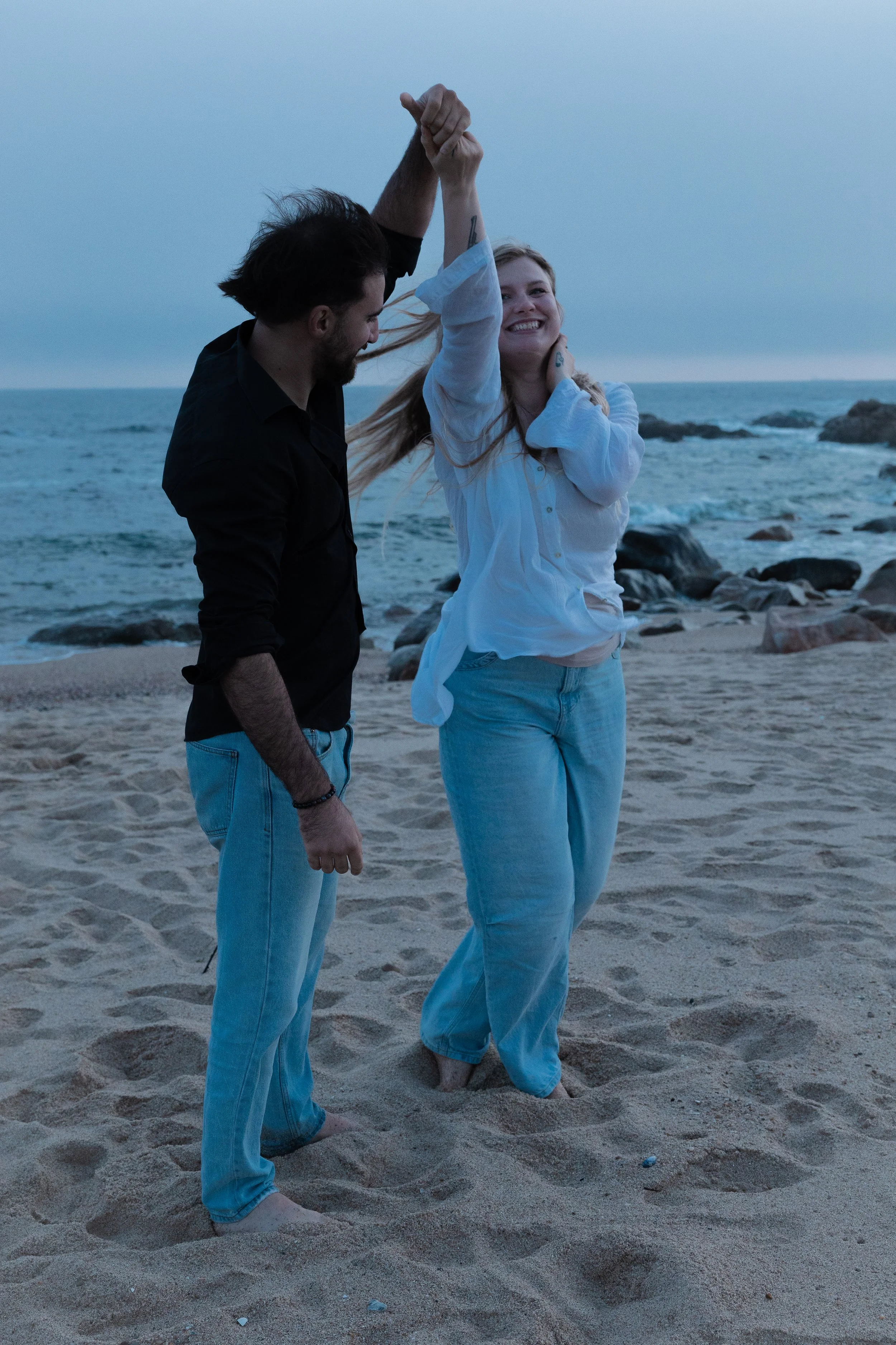 A young woman in a white shirt and blue jeans dancing with a man in a black shirt and blue jeans on a beach with rocks and ocean in the background during dusk.