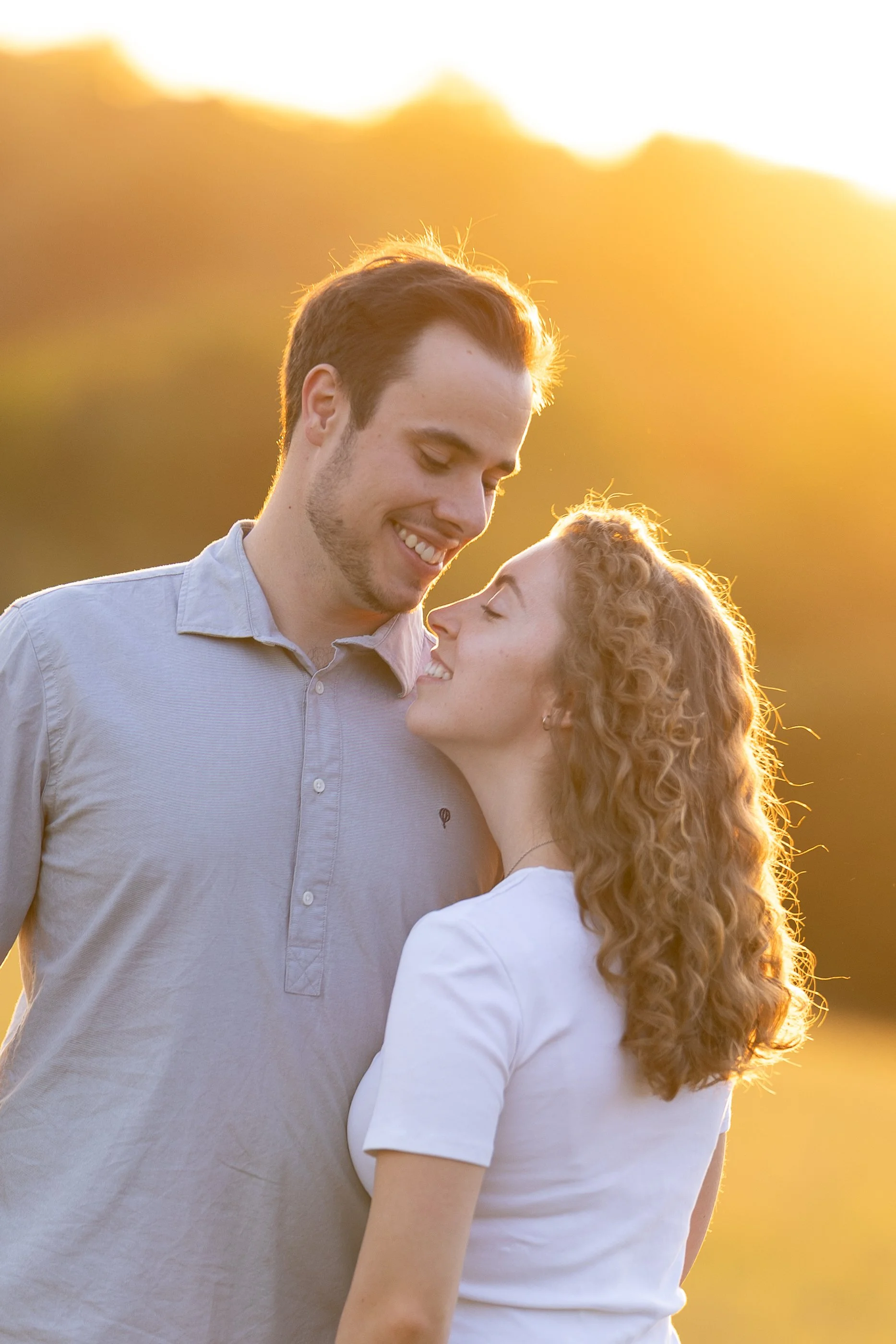 A young man with short dark hair and a young woman with curly blonde hair smiling and looking at each other outdoors during sunset.
