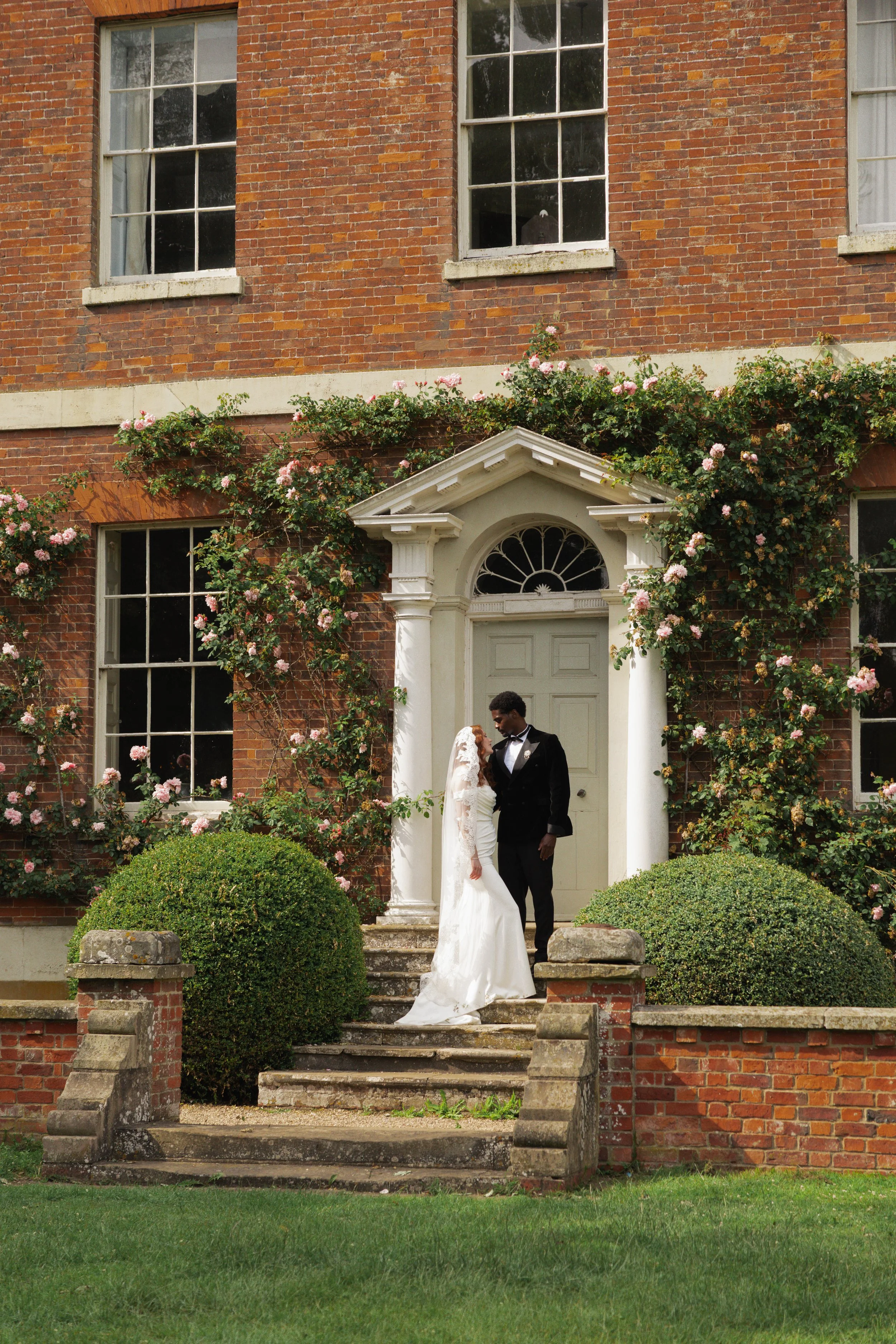 A bride and groom standing on the steps of a brick house with white columns and pink roses climbing on the facade. The bride is wearing a white wedding dress with a train and veil, and the groom is in a black tuxedo. They are looking at each other lovingly.
