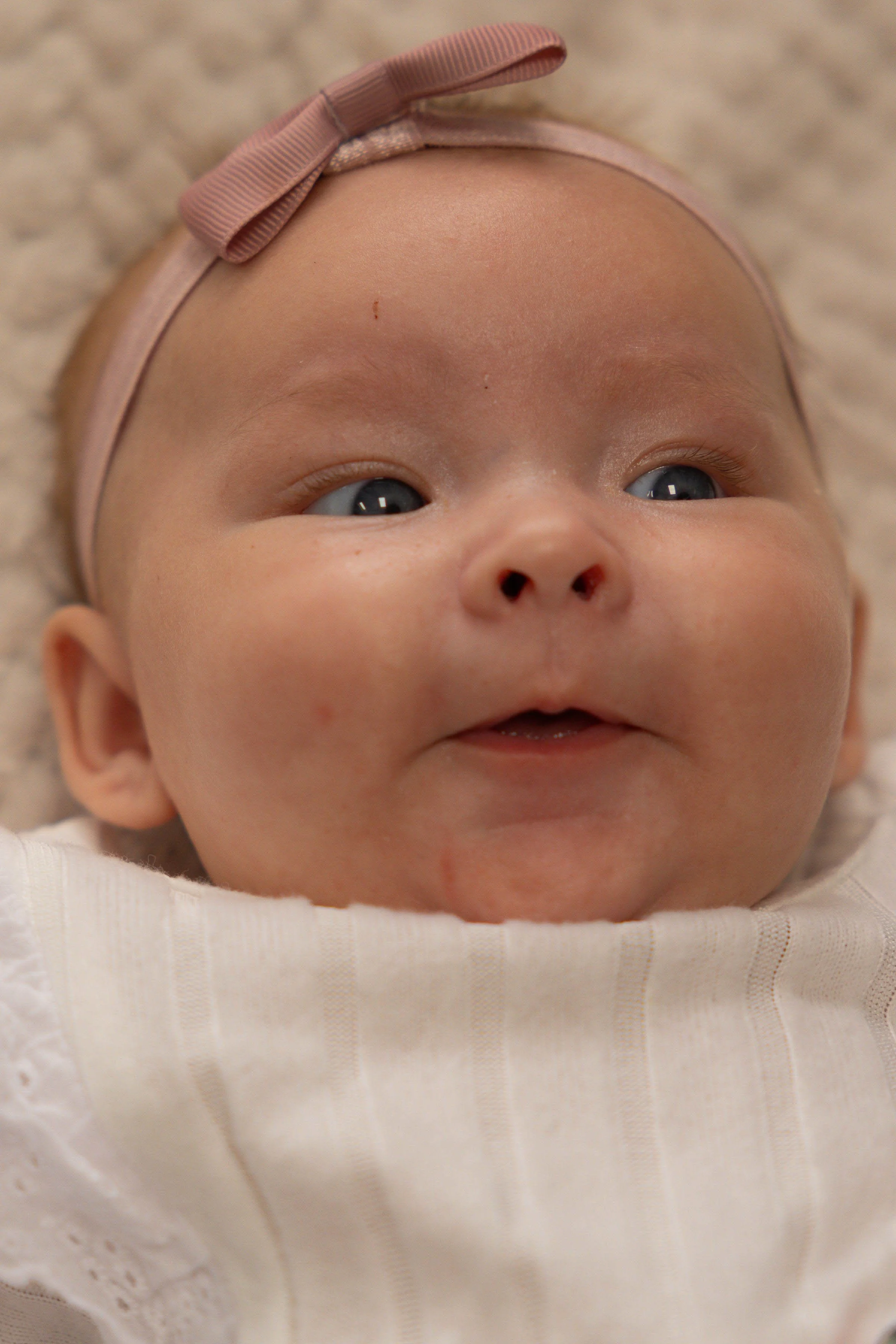 Close-up of a baby lying on a soft, textured beige surface, wearing a pink headband with a bow and a cream-colored top with a ribbed pattern.