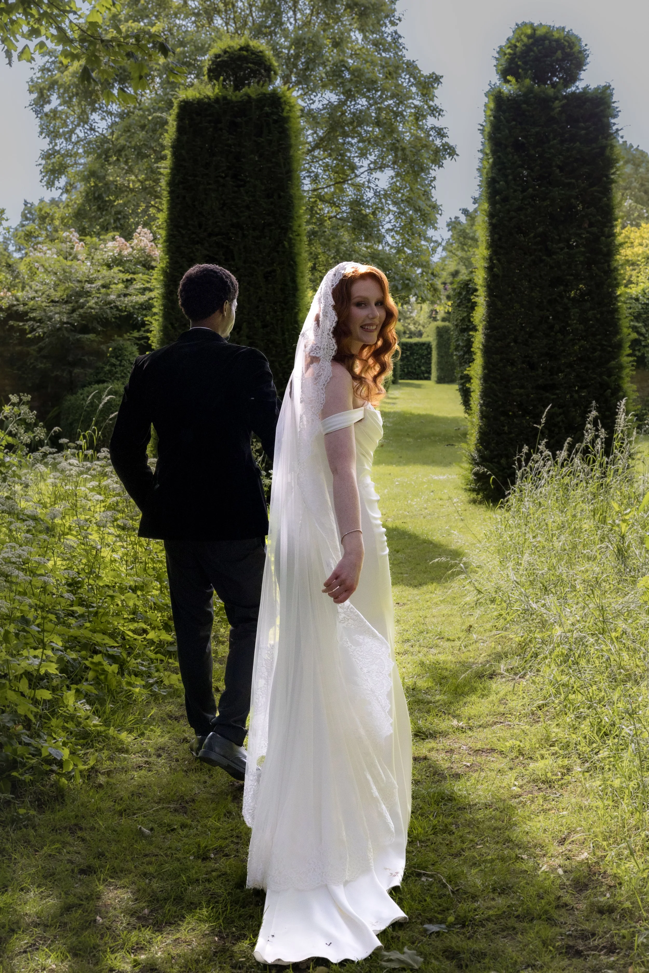 A bride with red hair in a white wedding dress and veil stands outside in a garden with greenery and large topiary trees, smiling while looking back at the camera. A man in a black suit stands nearby with his back to the camera.