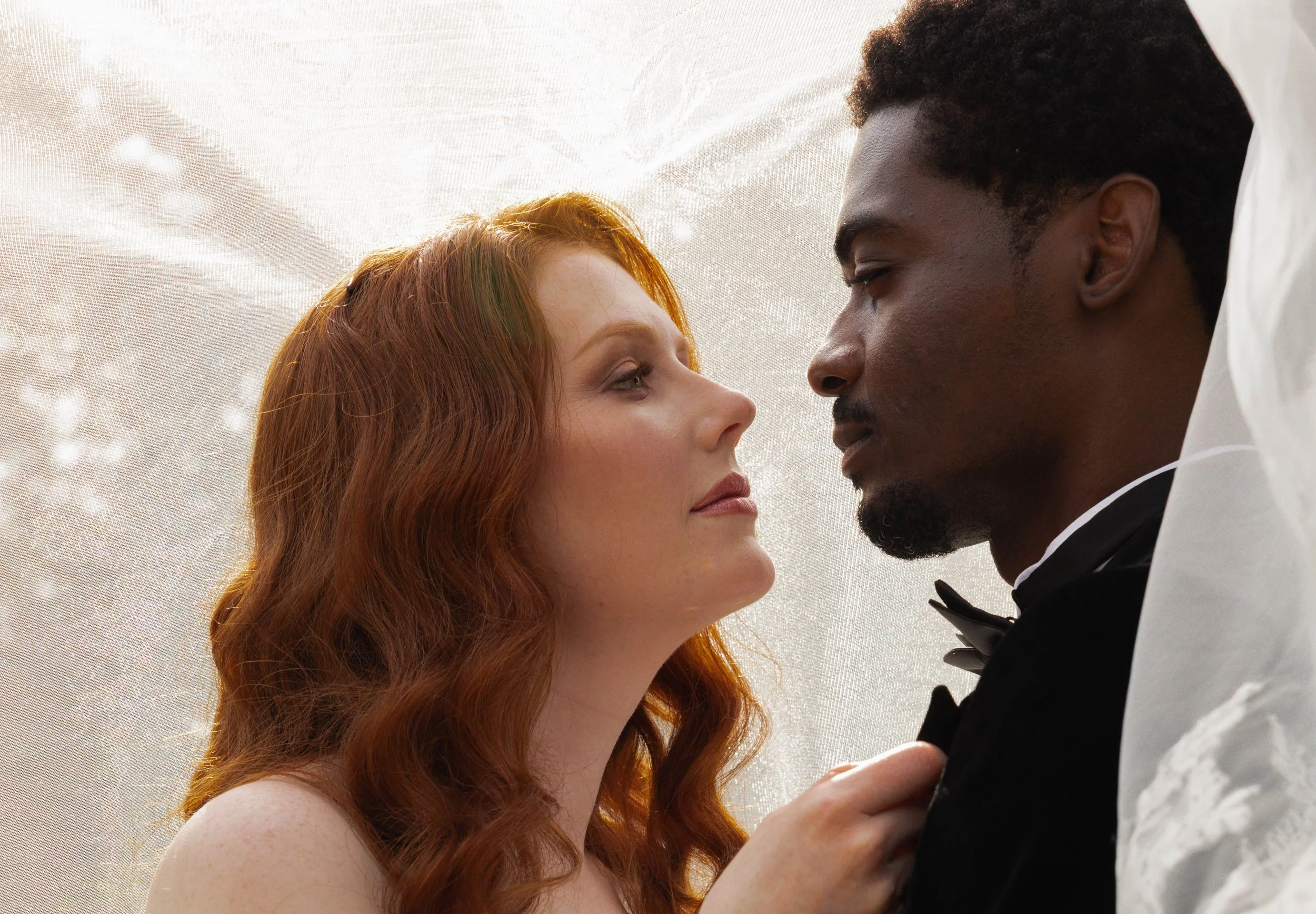 A couple dressed formally, sharing an intimate moment closely facing each other with their foreheads nearly touching, against a soft, light background.
