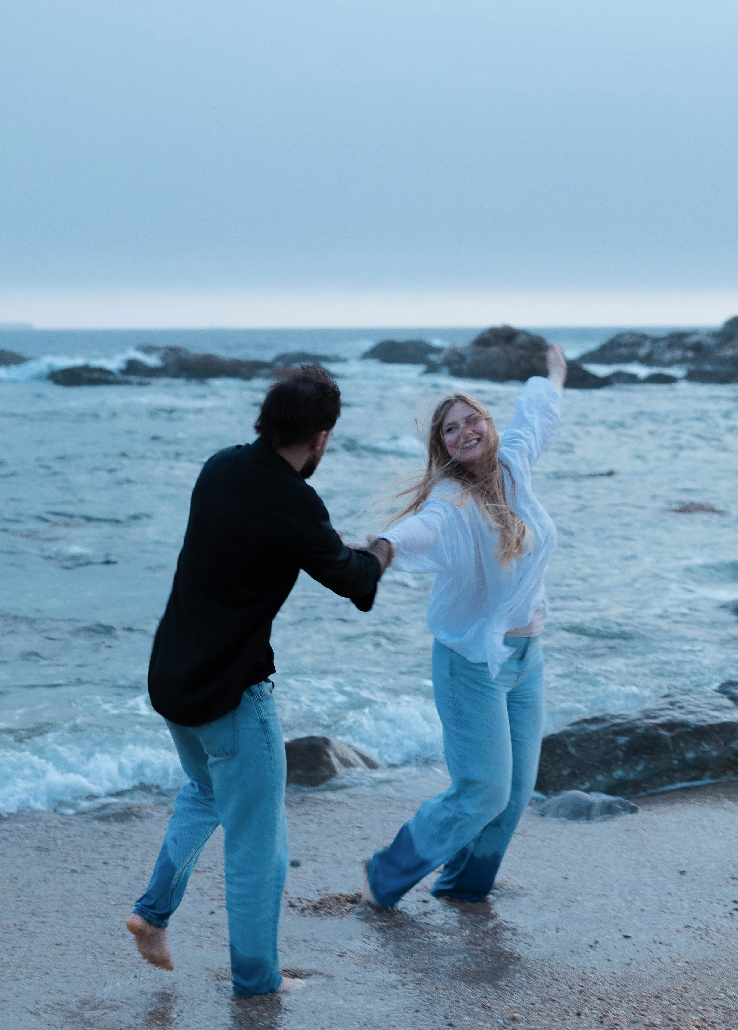 A smiling young woman in a white shirt and blue jeans holding hands with a man in a black shirt and blue jeans on a beach with ocean waves and rocks in the background.