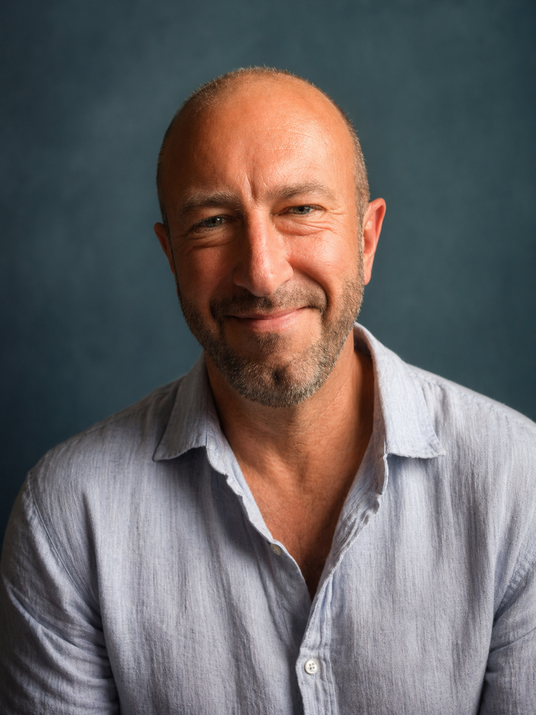 Close-up of a middle-aged man with a friendly smile, bald head, and gray beard, wearing a light-colored button-up shirt, against a dark teal background.