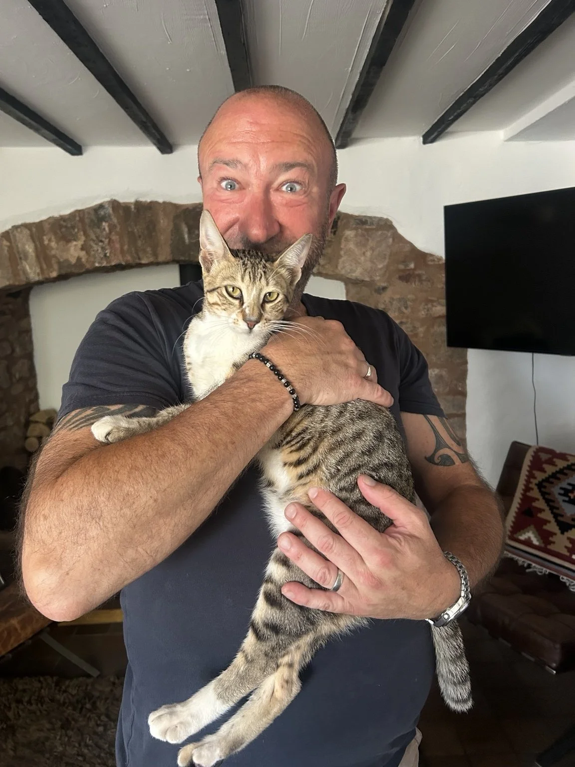 A man holding a tabby cat indoors, with a brick fireplace and a TV in the background.