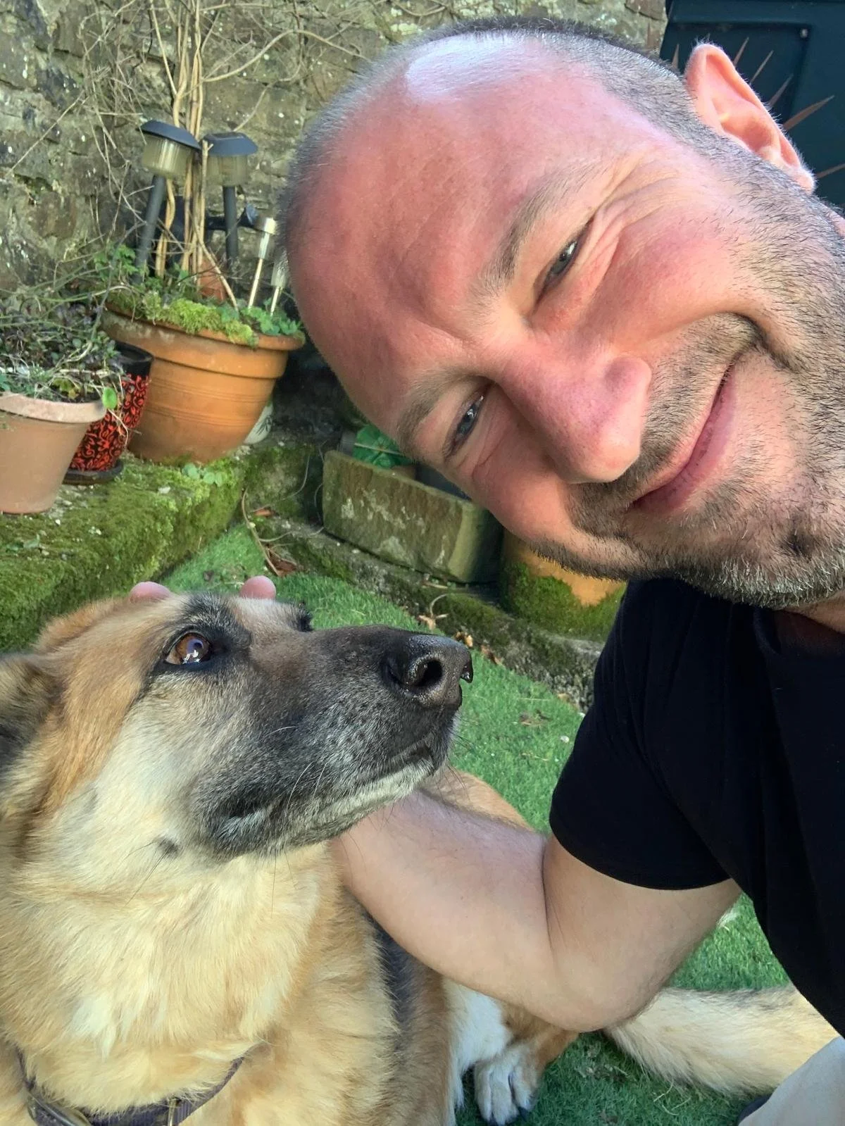 A man taking a selfie outdoors with his head turned to the side, smiling, next to a dog lying on the grass. The background includes potted plants and a stone wall.