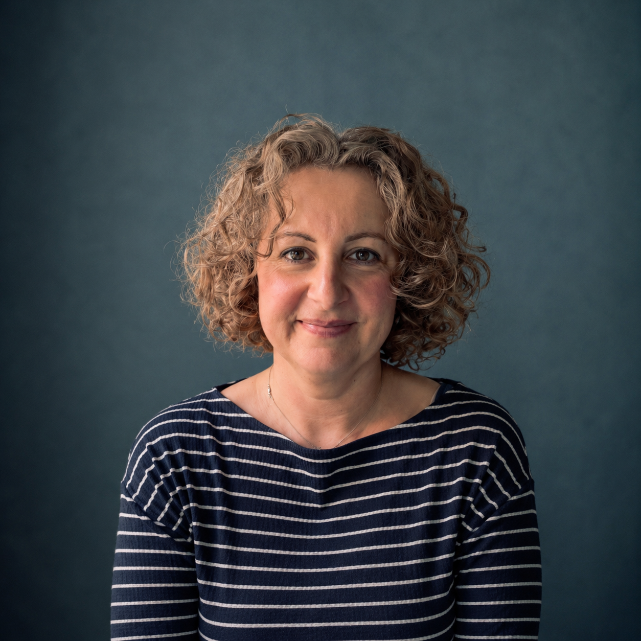 A woman with curly blonde hair wearing a navy and white striped shirt, smiling softly against a dark teal background.