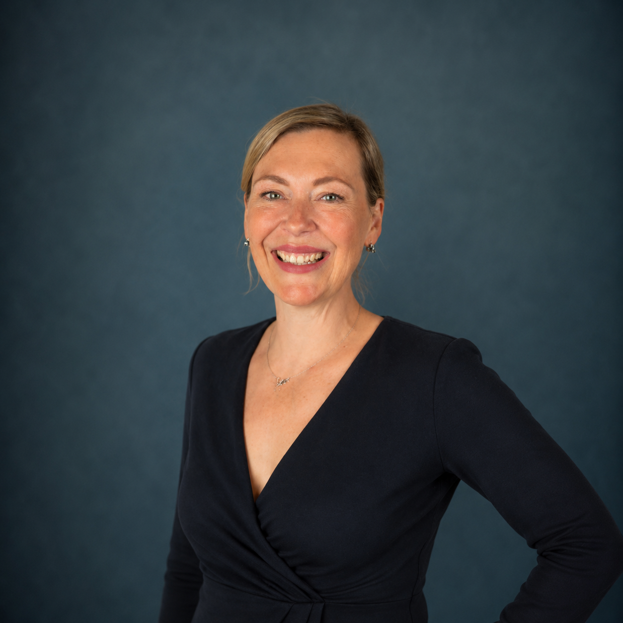 A smiling woman with short blonde hair, wearing a black dress and jewelry, standing against a dark teal background.