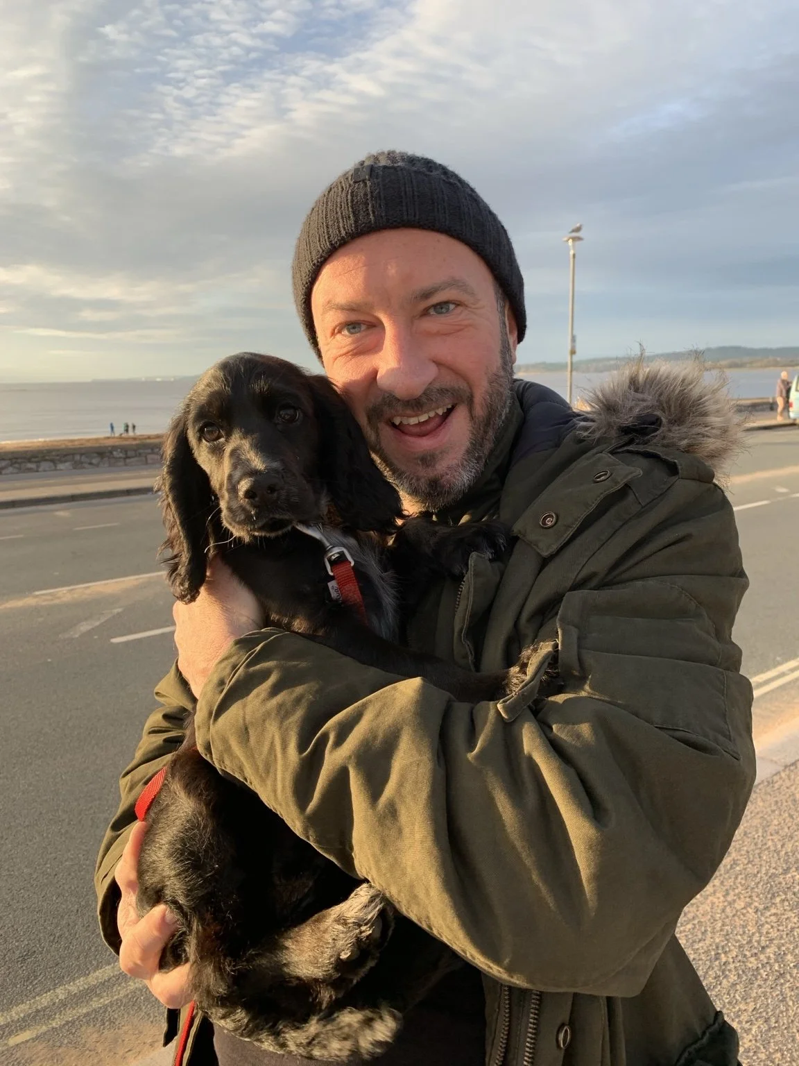 A man wearing a black knit hat and dark green jacket holding a black and tan puppy with floppy ears, near a body of water and a cloudy sky.
