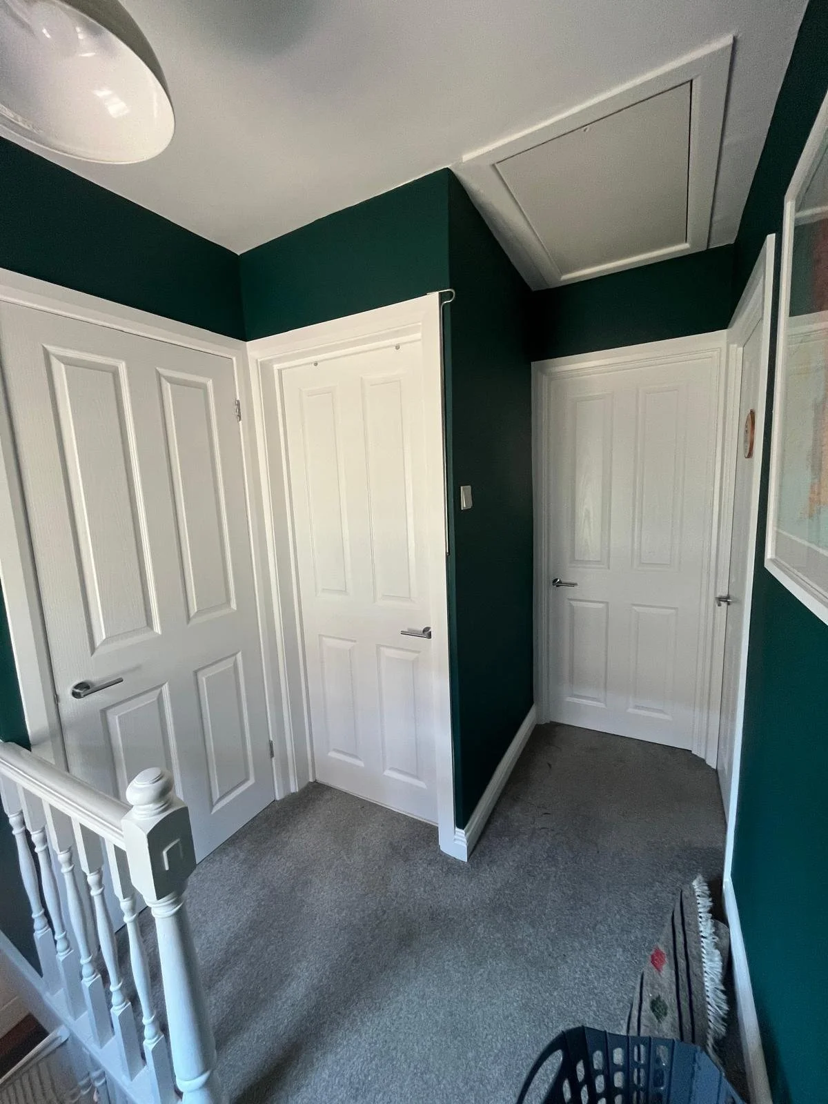 A small hallway with dark green walls and three white doors, one of which is in a closet or attic access space in the ceiling. The hallway has gray carpet and a white staircase railing on the left, with a small black basket on the floor. There is also a painting or framed picture on the wall to the right.