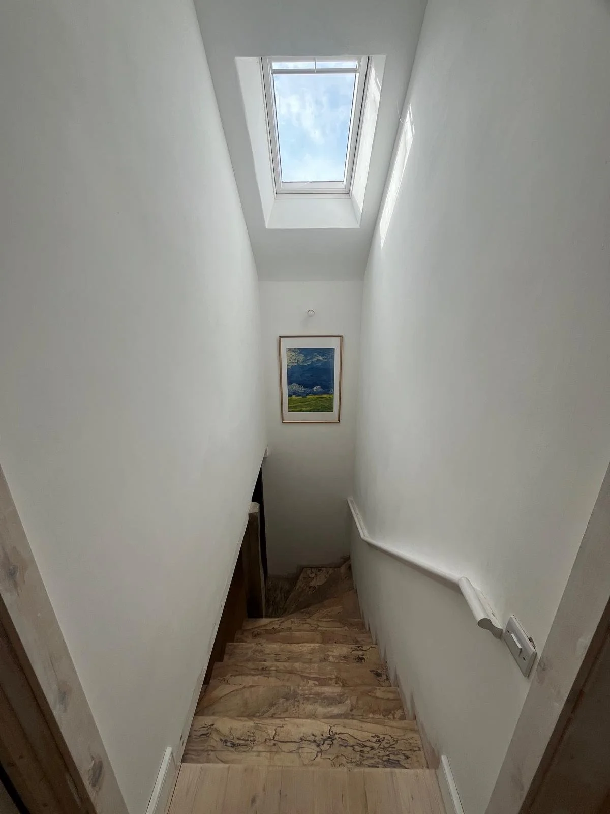 View of a narrow staircase with wooden steps, white walls, a staircase handrail on the right, a ceiling window allowing natural light, and a framed landscape painting on the wall ahead.