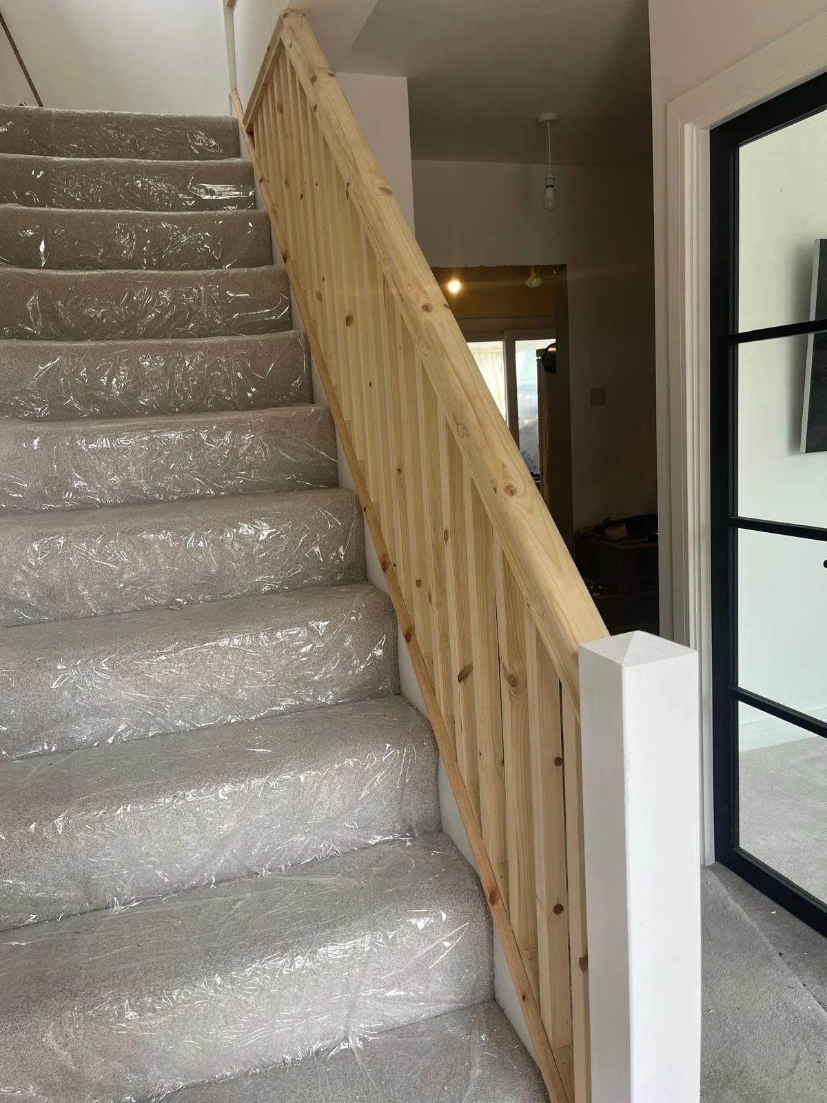 Interior view of a staircase with new wooden railing and protective plastic covering on carpeted stairs, next to a glass-paneled door.