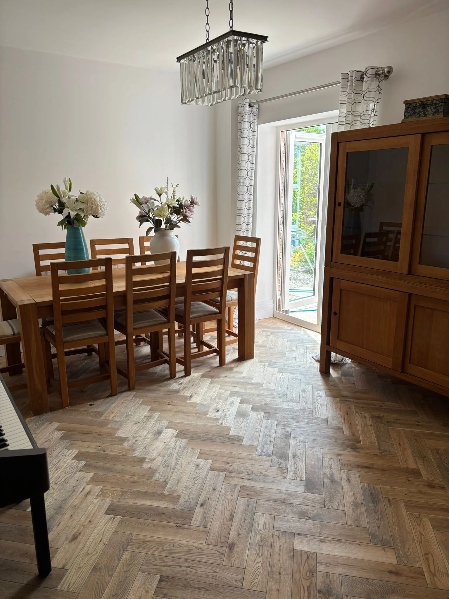 Dining room with a wooden dining table, six matching chairs, two vases with white and pink flowers, a wooden cabinet, open door leading outside with trees, white curtains, and a modern chandelier hanging from the ceiling.