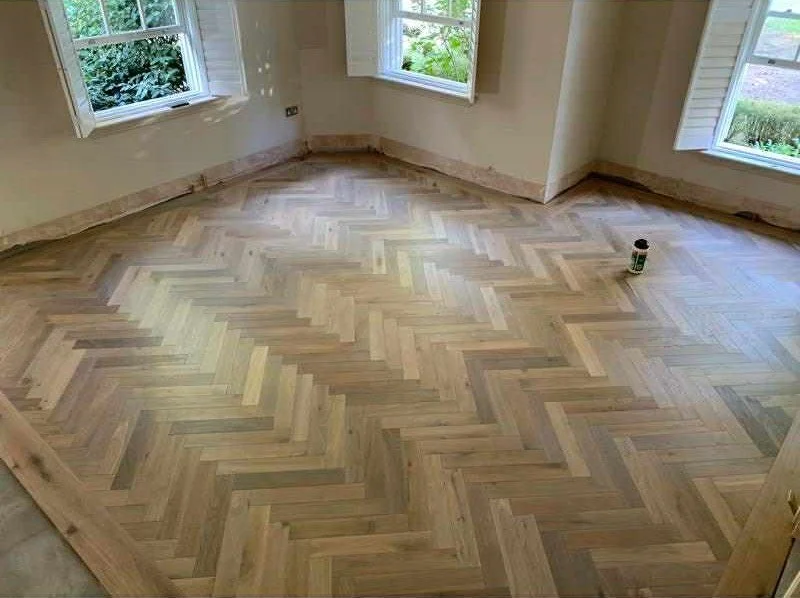 A room with newly installed herringbone wood flooring, three windows, and a small container of finish or stain on the floor.