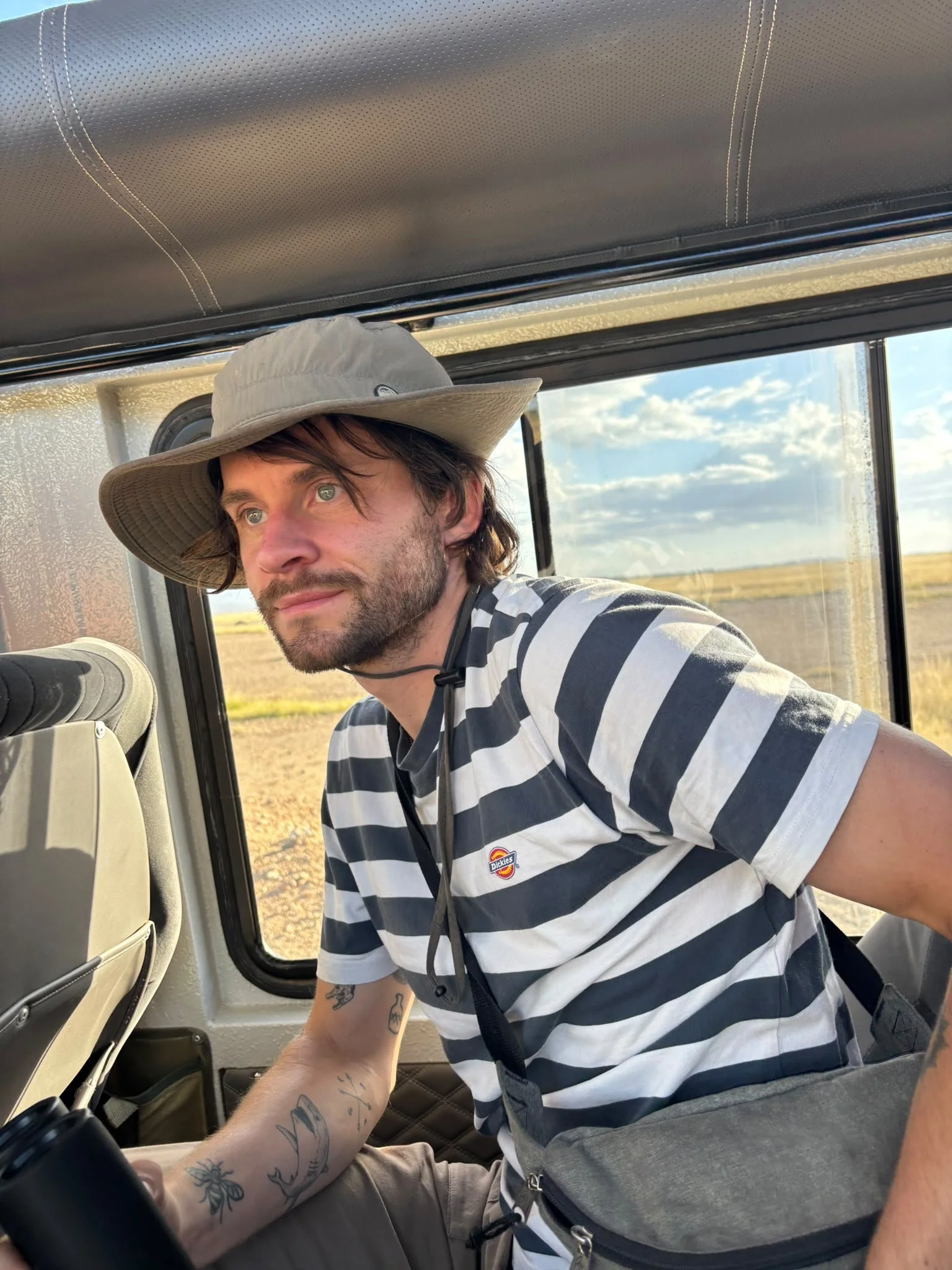 A man with tattoos on his arm, wearing a striped T-shirt and a wide-brimmed hat, is sitting inside a vehicle with a window showing a rural landscape with a partly cloudy sky in the background.