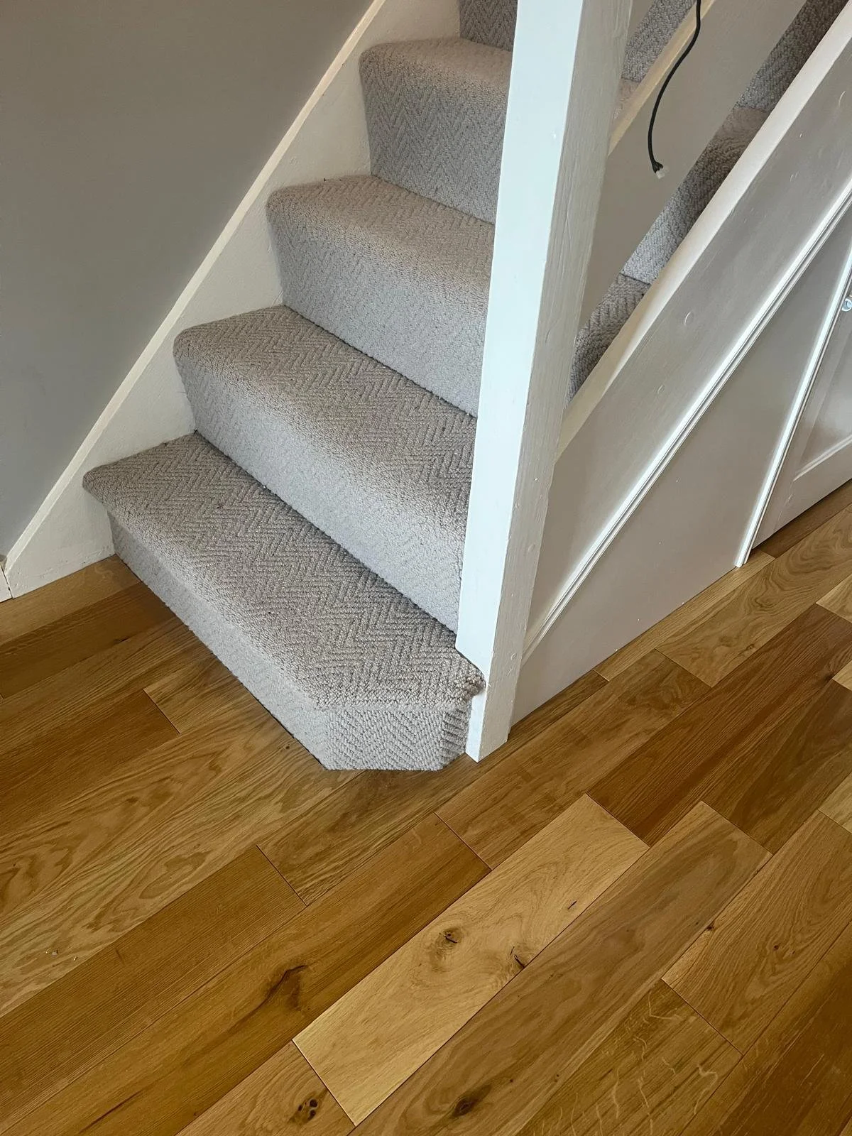 Carpeted stairs with beige textured fabric, next to a section of beige wall and a white vertical panel, with hardwood flooring nearby.