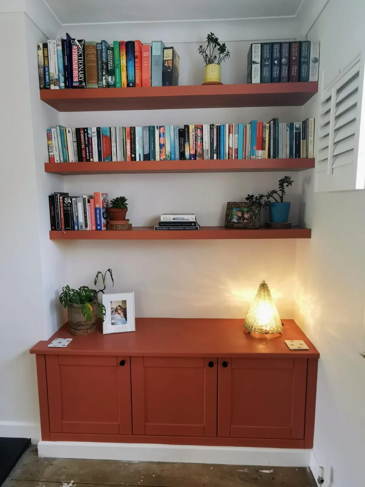 A white wall with three peach-colored wall-mounted shelves filled with books and decorative plants. Below the shelves is a peach-colored cabinet with two doors, a potted plant, a framed photo, and a lamp emitting warm light.