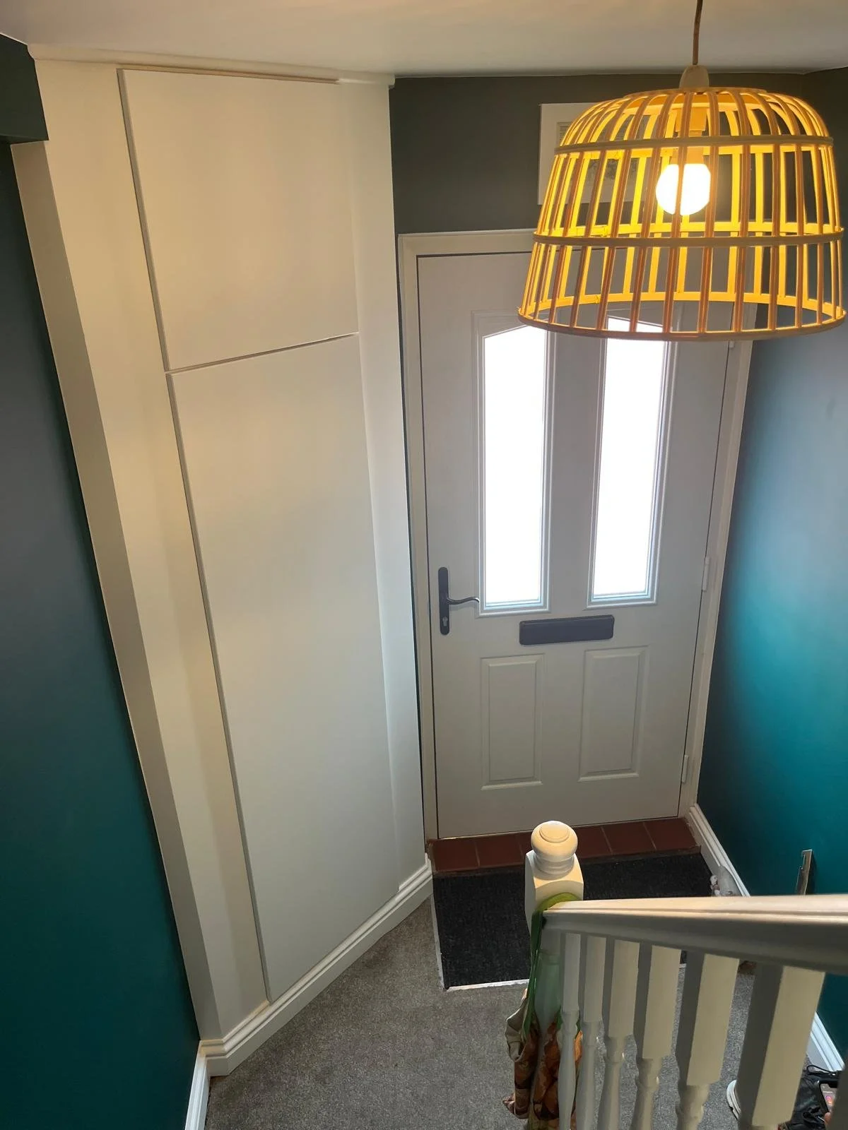 An entryway with a white door that has two narrow glass panels and a mail slot, a teal wall, a large white storage cabinet, a wooden hanging light fixture, and a staircase with a white railing at the bottom.