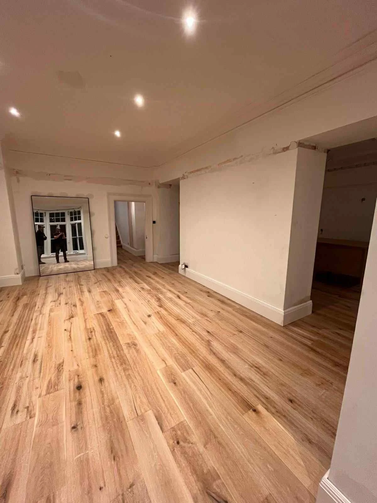 An empty living room with wooden floors, white walls, and ceiling, featuring recessed lighting. A large mirror is leaning against the wall, reflecting two people taking the photo. There is a doorway and a window in the background.