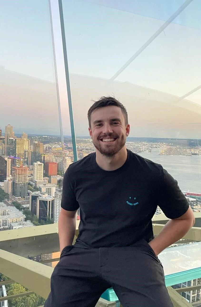 A man smiling while sitting on a glass observation deck high above a city skyline with a river in the background.