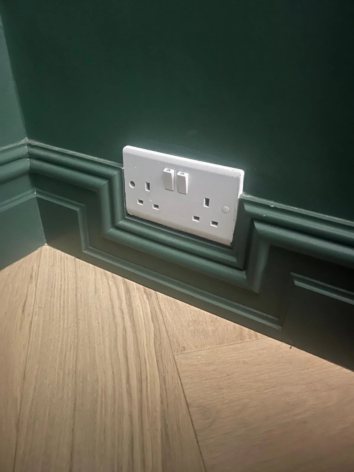 A white electrical outlet with two switches on a green wall near a wooden floor.