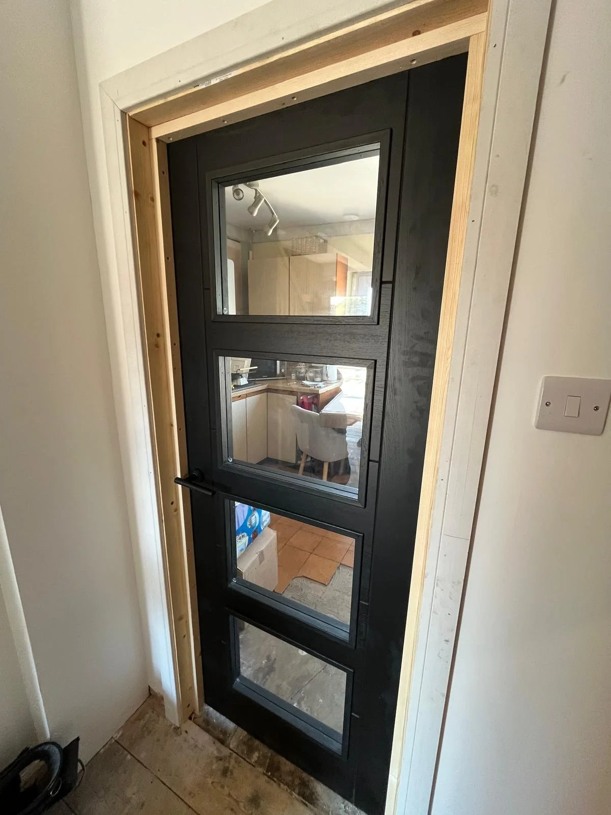A black door with four glass panels, leaning against a wall in a home kitchen, with a light switch on the wall to the right.