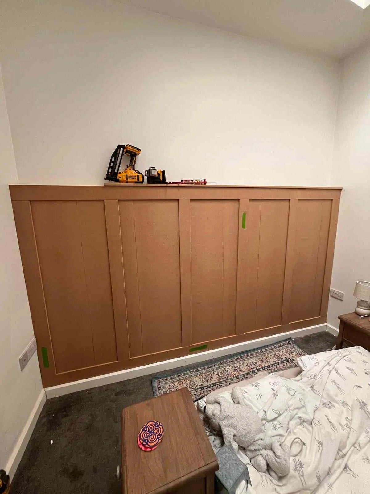 Room with wooden headboard or divider, tools on top, bed with torn sheets, small nightstand with lamp, and a colorful coaster on a wooden table.