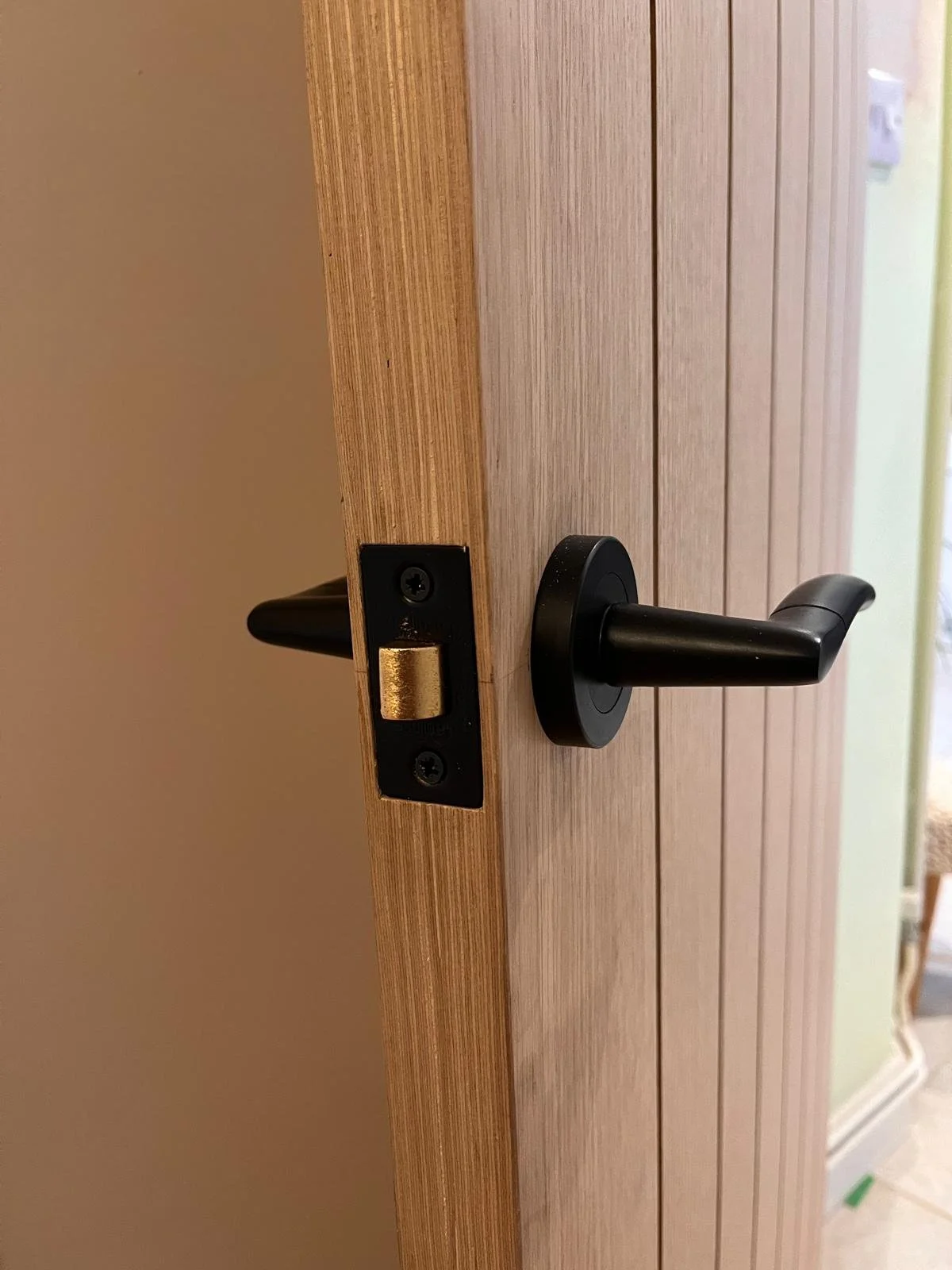 A door partially open showing its latch and handle, with a wood grain design on the door and a light-colored wall in the background.