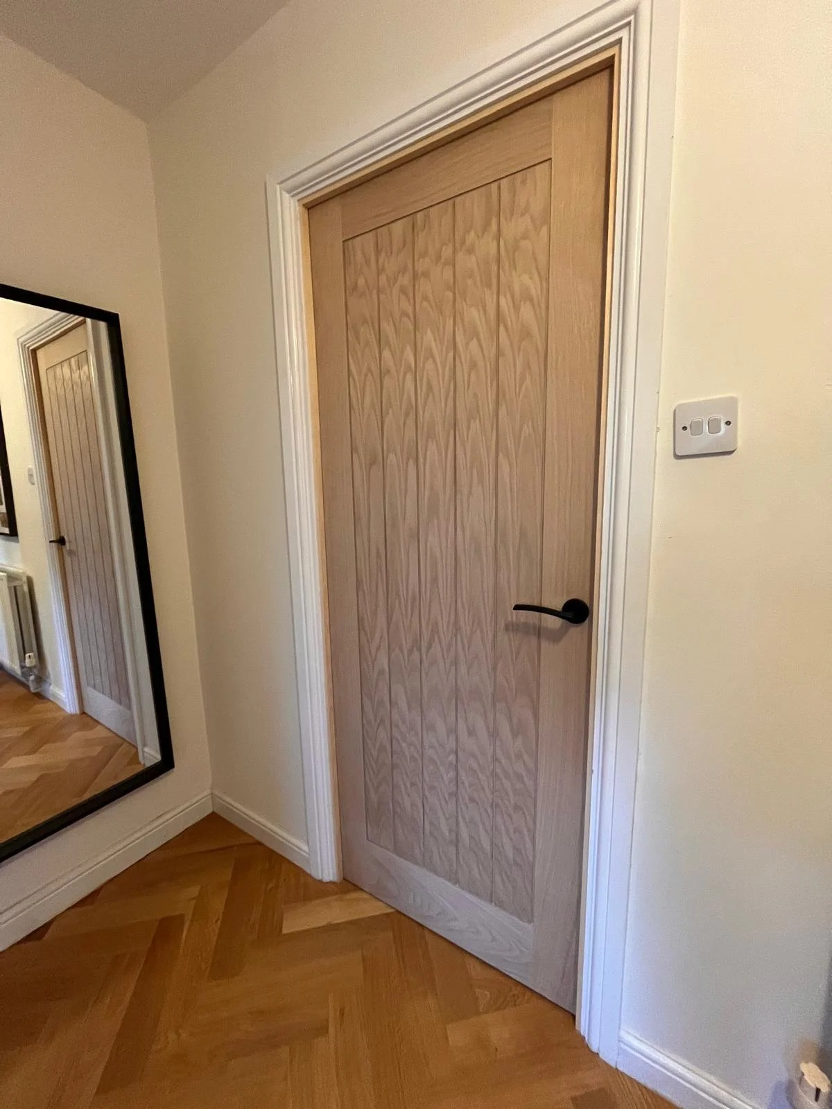 A wooden interior door with a black handle is in a room with beige walls and a wooden parquet floor. A mirror and a white door frame are visible on the adjacent wall.