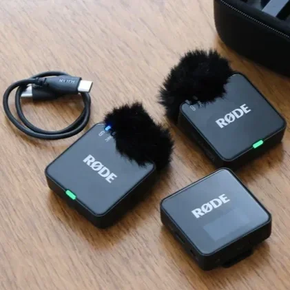 Three Rode wireless microphones on a wooden surface, with one microphone connected to a charger and two with furry windshields.