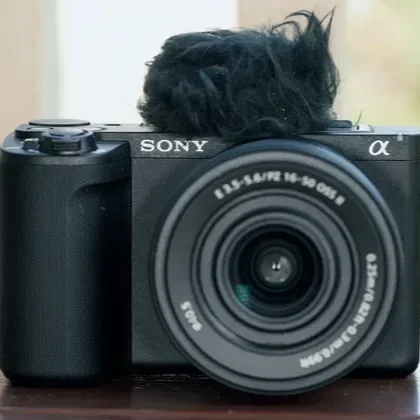 A Sony mirrorless camera with a large lens, placed on a surface with a fluffy black object and a blurred background.