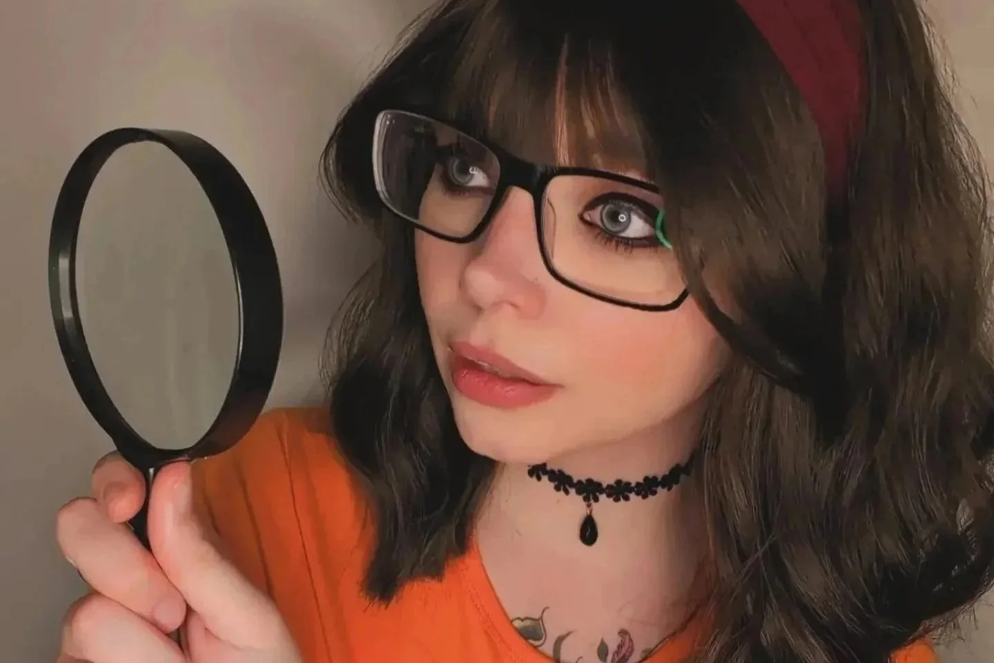 A woman with dark, wavy hair, wearing glasses, an orange top, and a black choker necklace, holds a mirror and looks at her reflection.