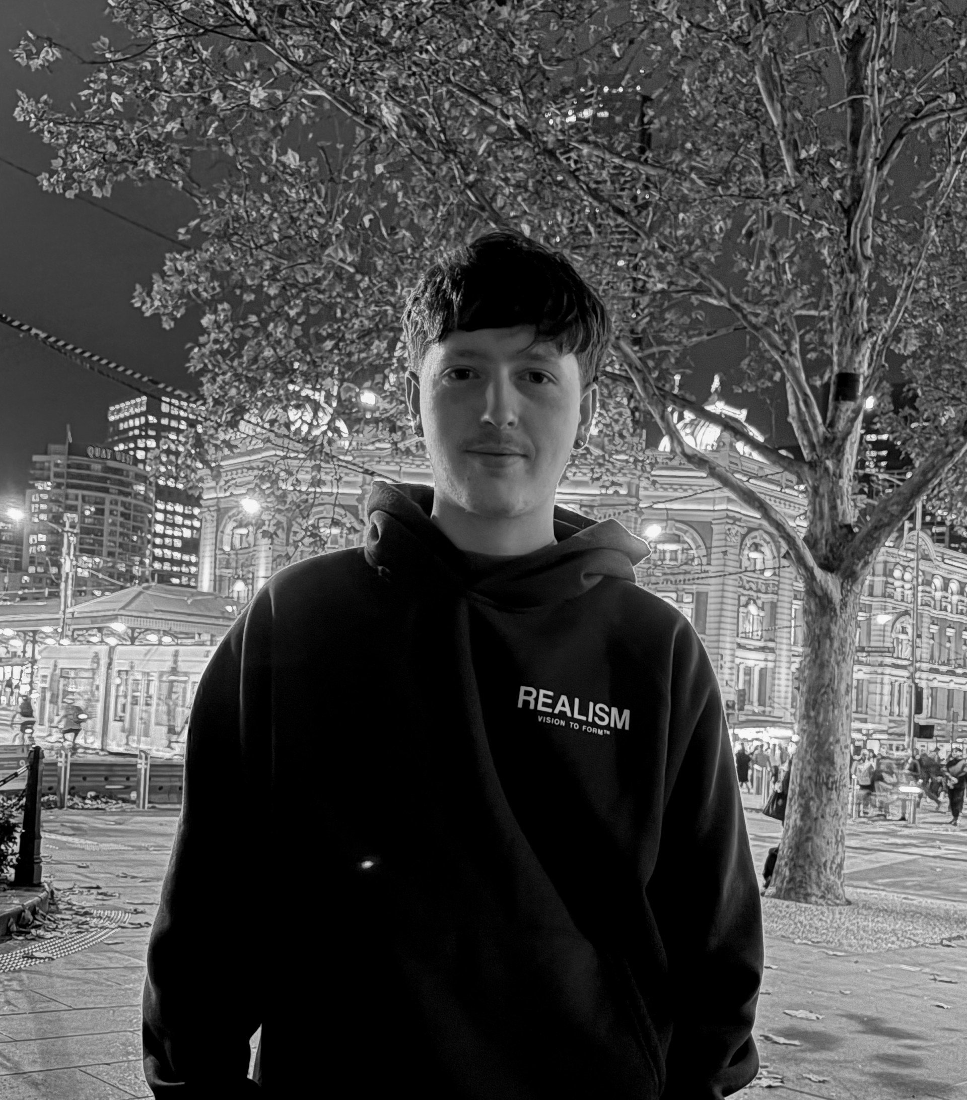 A young man with short dark hair is standing outdoors at night in front of a large tree with many leaves, illuminated urban buildings, and a carousel with lights, all in black and white.