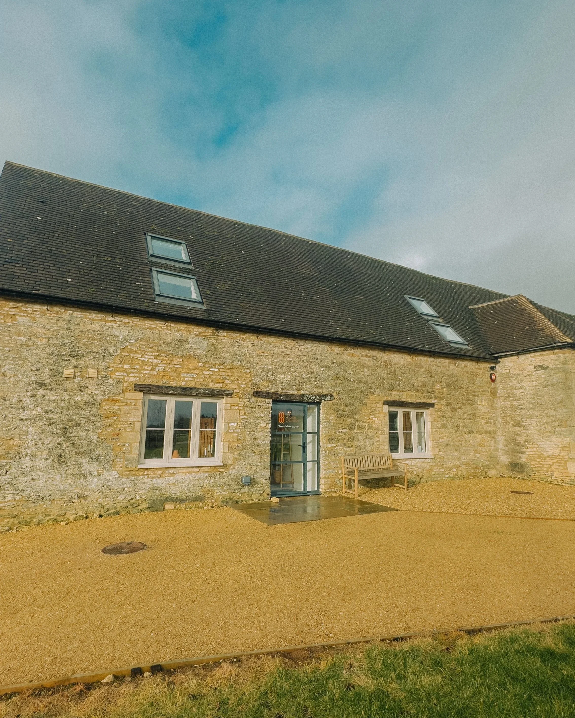 A stone building with three skylights on the roof and two large windows on the front, a door between the windows, a wooden bench outside, and a gravel area in front with patches of grass and a drain.