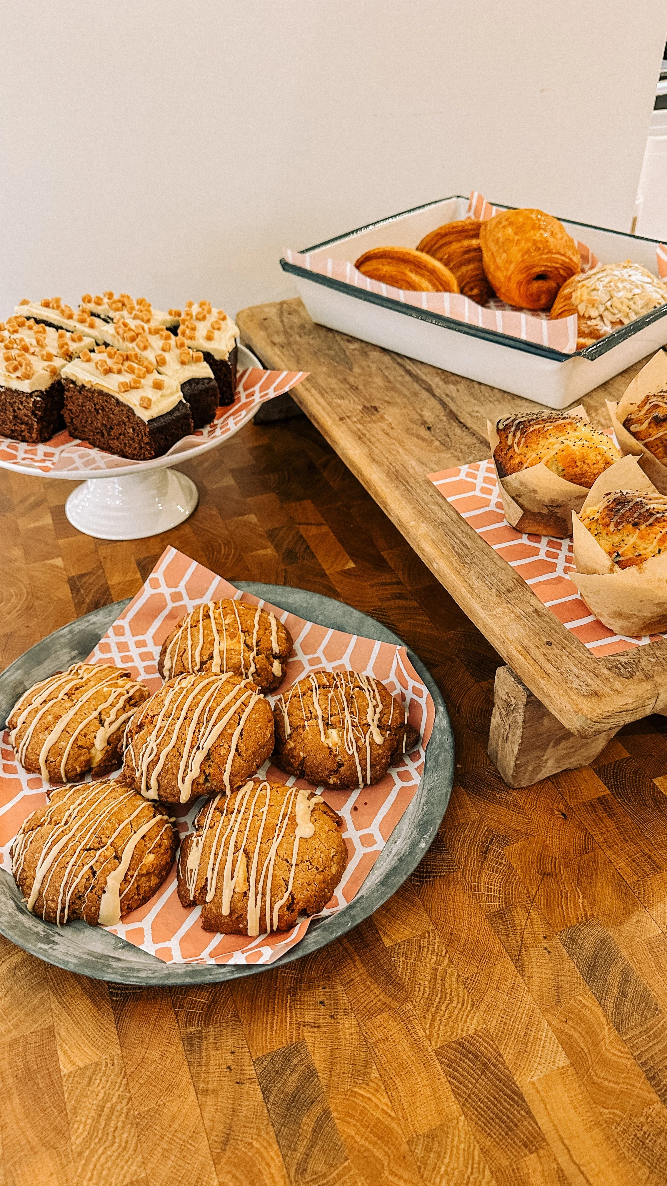 Assorted baked goods, including cookies, croissants, muffins, and slices of cake, displayed on various trays and stands on a wooden table.