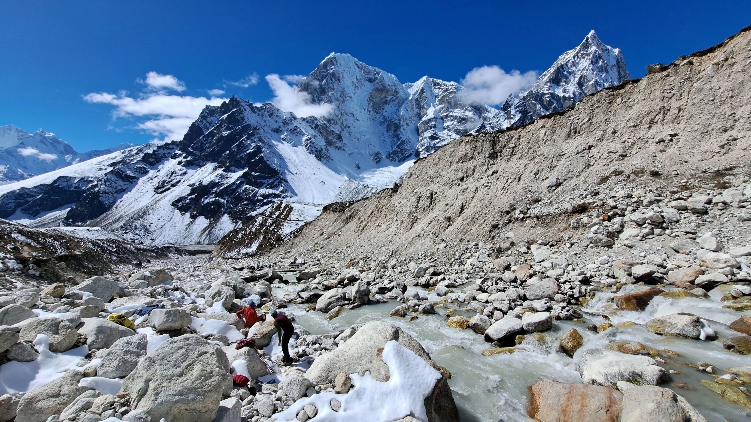 Mountain landscape with snow-capped peaks, rocky foreground, and a river with people crossing it.