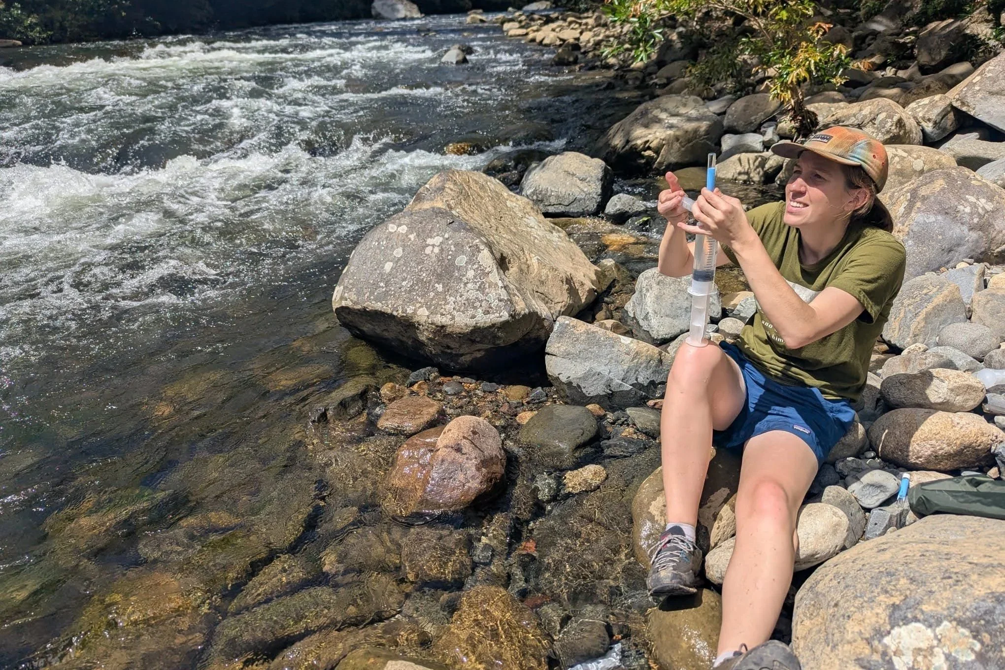 A woman sitting on rocks by a flowing stream, holding a syringe, wearing a hat, green T-shirt, and blue shorts, smiling and enjoying the outdoor setting.
