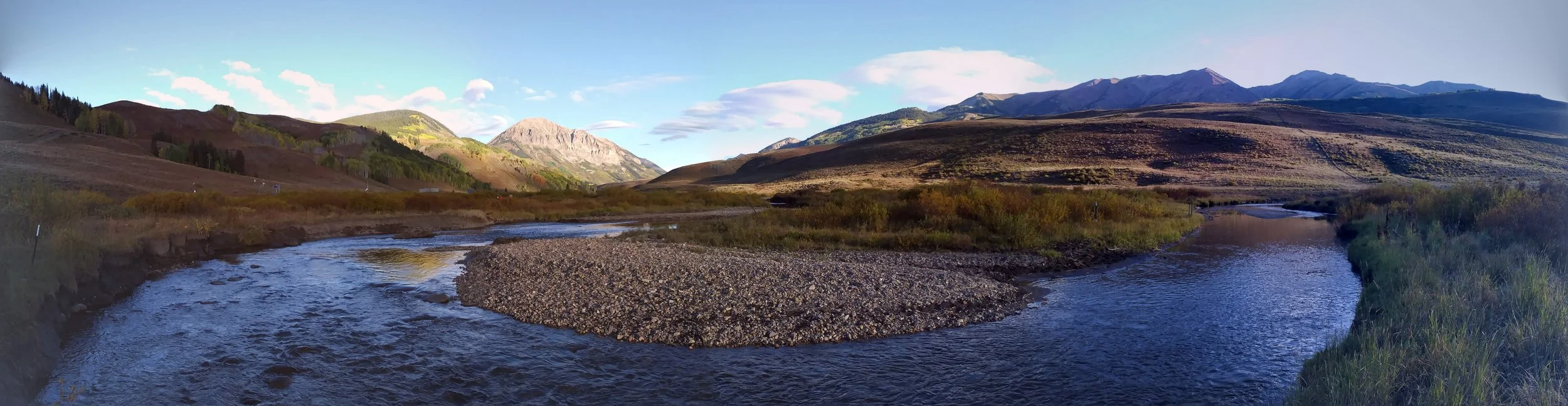 Panoramic view of a river flowing through a valley with mountains in the background, under a partly cloudy sky.