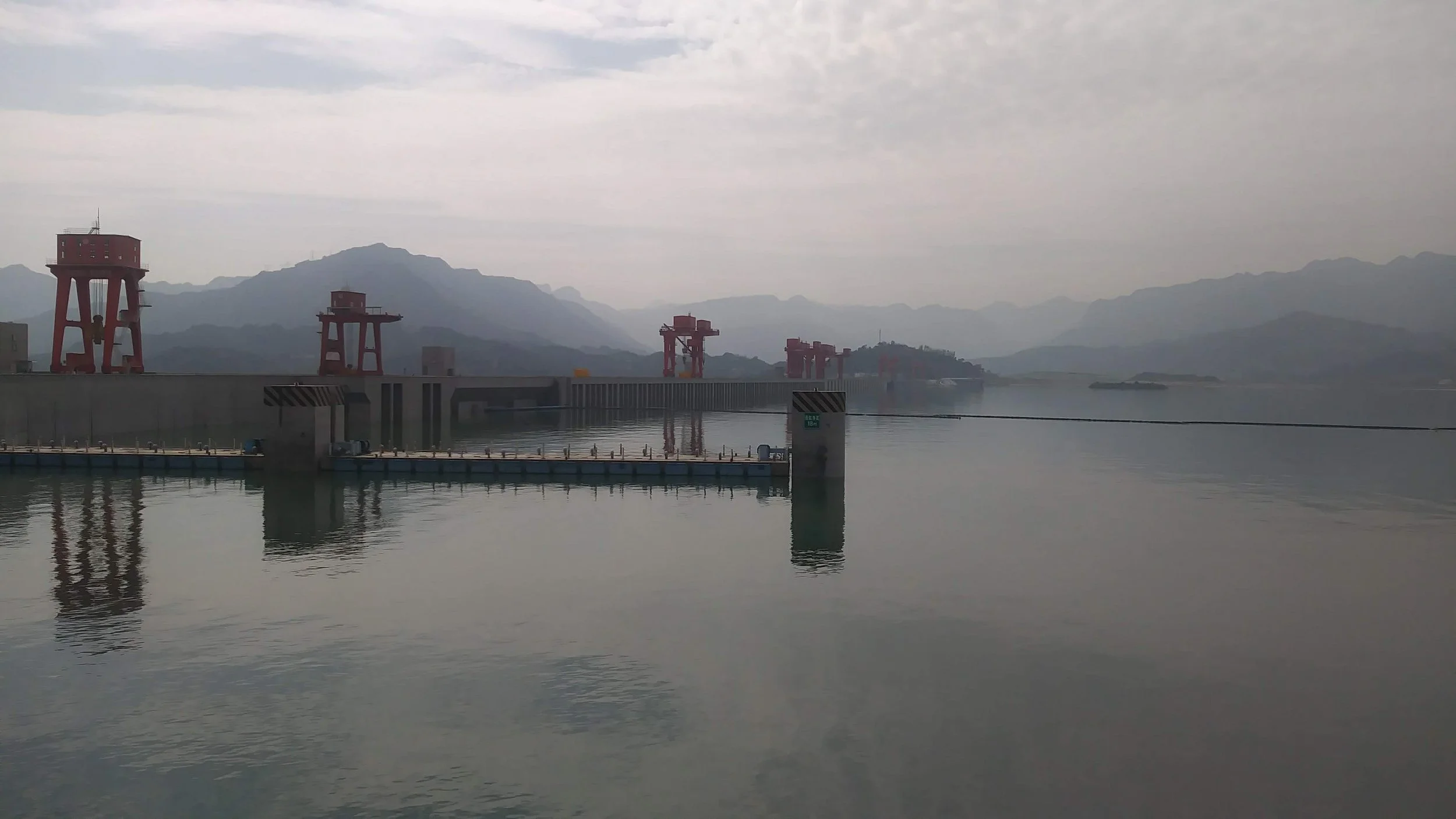 A large body of water with a dock and red cranes along the edge. Mountain ranges and a cloudy sky are visible in the background.