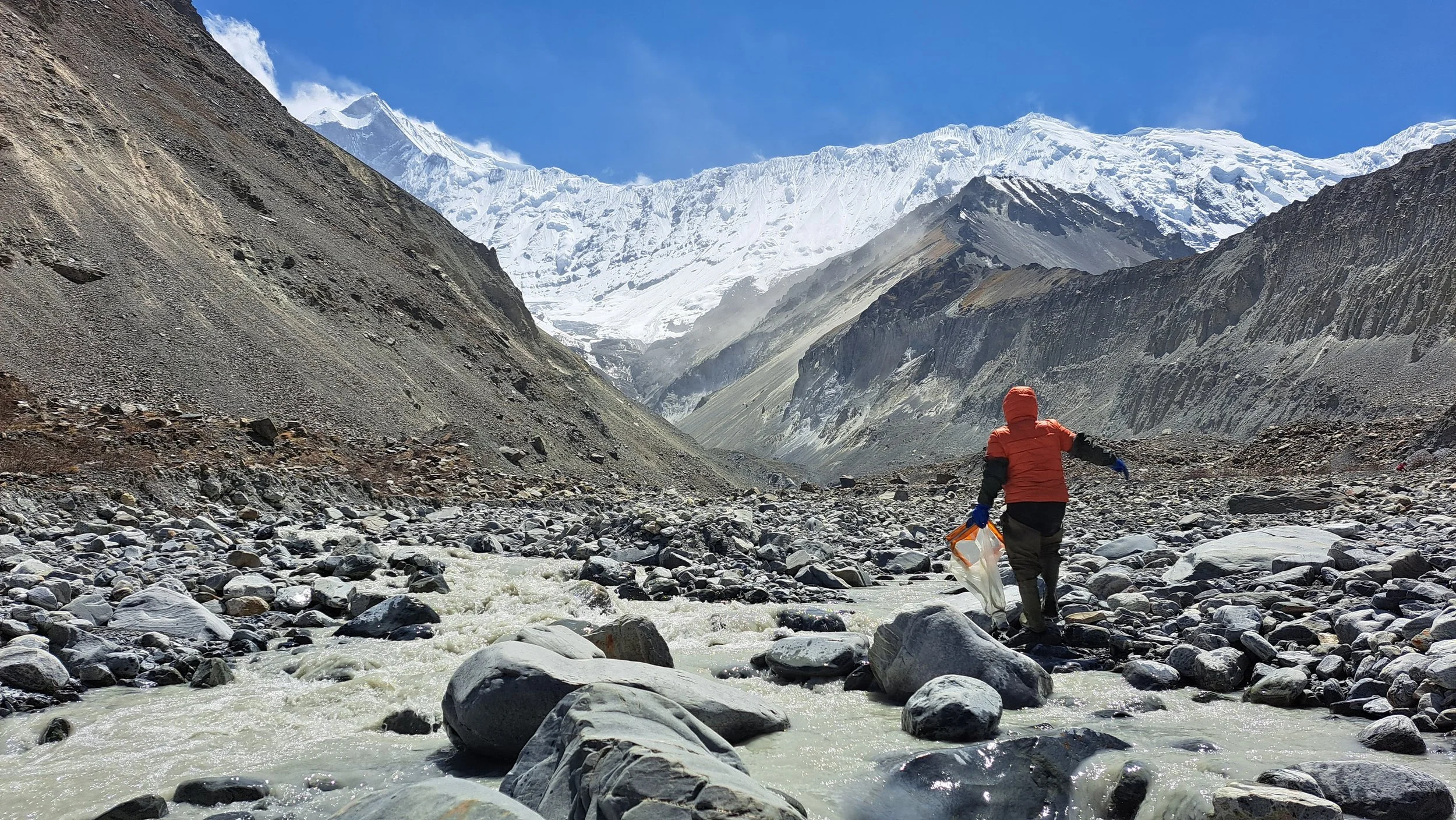 A person dressed in an orange jacket and black pants, carrying some items in a plastic bag, walking across rocky terrain with a river, set against a backdrop of towering snow-capped mountains and a blue sky.