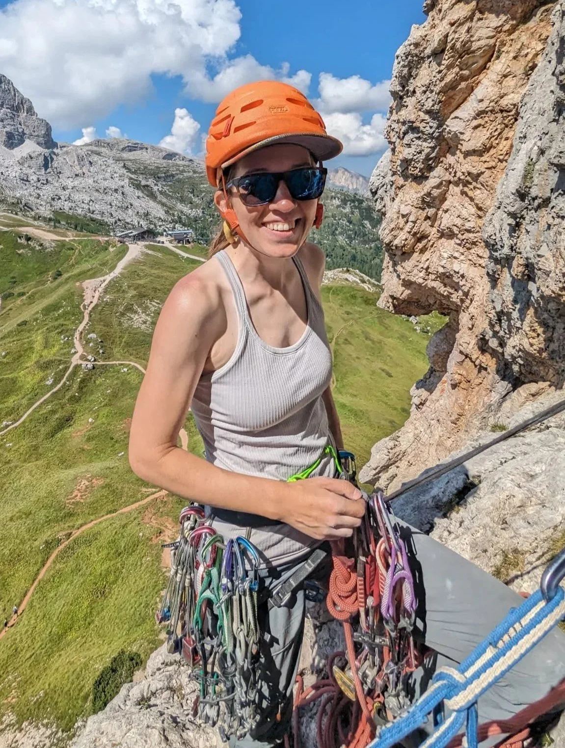 A woman rock climbing outdoors, wearing an orange helmet, sunglasses, a grey tank top, and climbing gear, with a mountainous landscape and blue sky in the background.