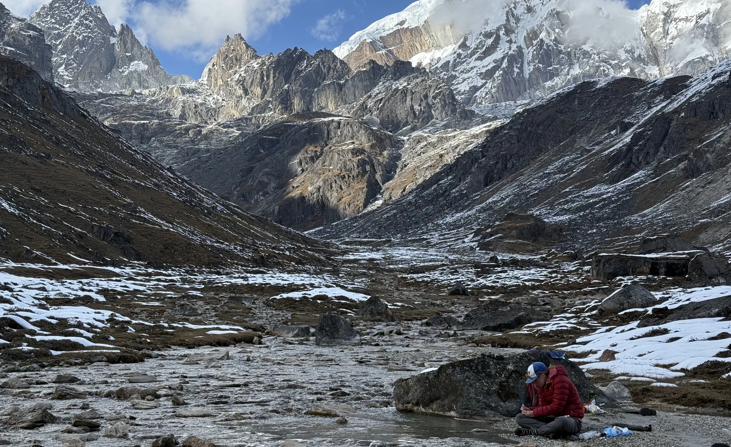 A mountainous landscape with snow-capped peaks, rocky slopes, and a valley with a flowing stream. Two hikers sit near the stream in the foreground, one wearing a red jacket and blue cap, surrounded by rocks and patches of snow.