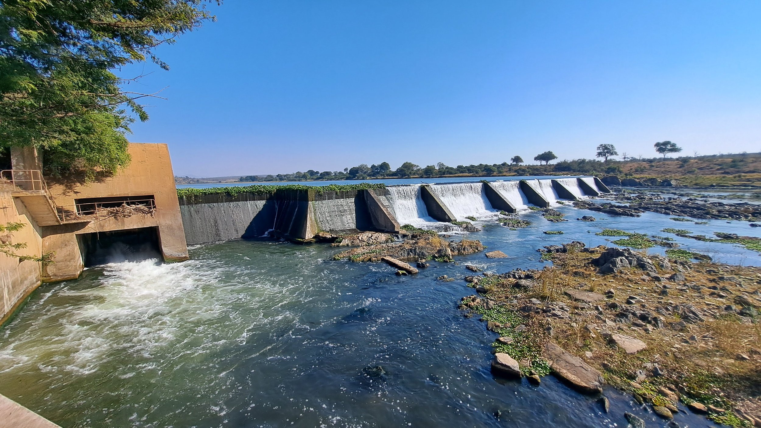 A dam with water flowing over its spillways, surrounded by a rocky riverbank and trees in the background, under a clear blue sky.