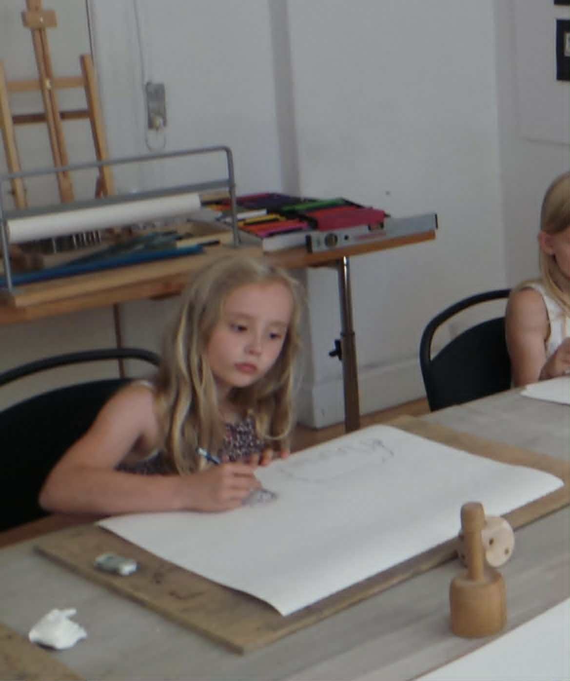 A young girl (Anna Ottilie Ravnsberg) with blonde hair sits at a table with a large sheet of paper in front of her. The setting appears to be a classroom or art room, with art supplies and tools visible on the table and shelves behind her.