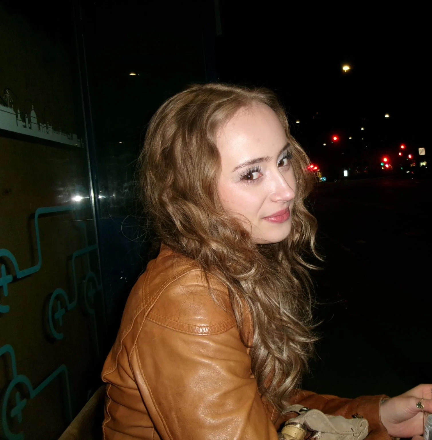 A young woman (Anna Ottilie Ravnsberg) with long, curly hair and light skin, wearing a brown leather jacket, looking over her shoulder at night. She is sitting outside a bus stop at night, with city street lights visible in the background.
