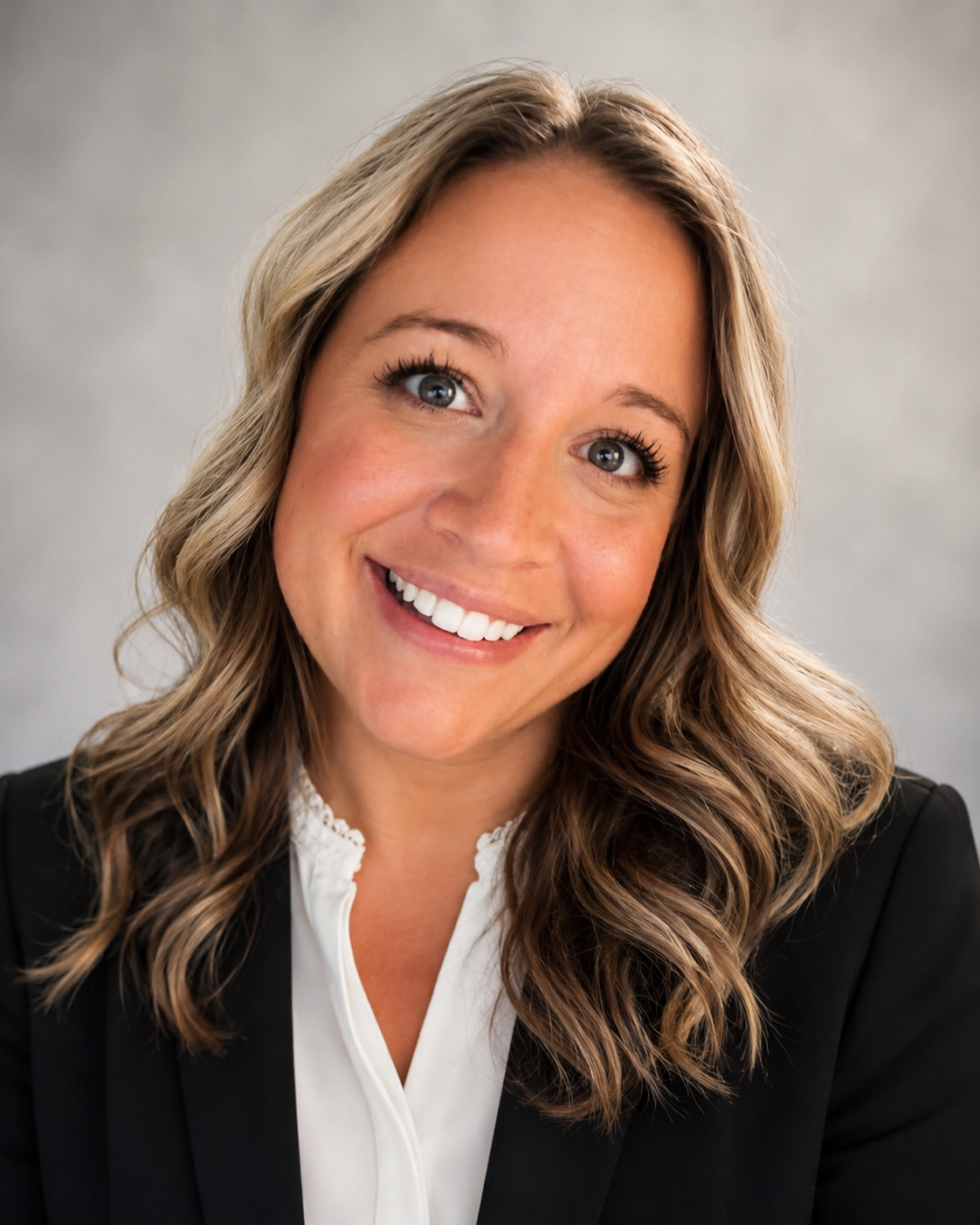 A smiling woman with wavy, shoulder-length blonde hair, wearing a black blazer and white blouse, against a neutral gray background.