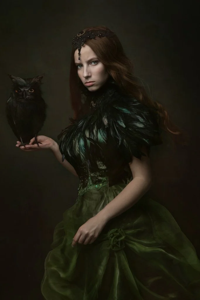 Een jonge vrouw met rood haar, in een donkere, elegante jurk met veren, houdt een zwarte uil op haar hand, achtergrond donker.