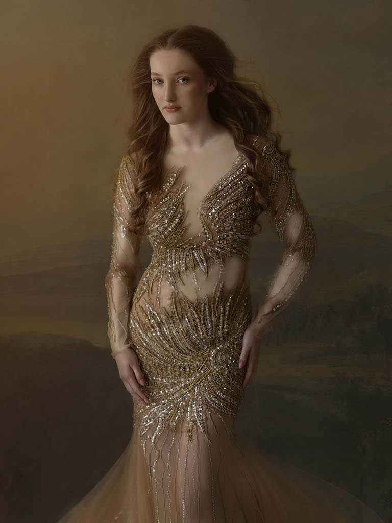 Vrouw in een goudkleurige, versierde jurk met lange mouwen en een uitgesneden decolleté, poseert voor een gele achtergrond.